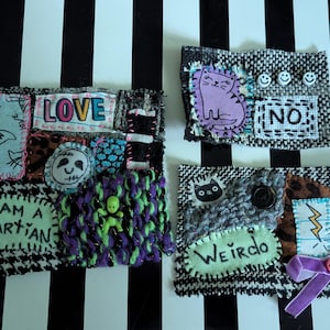 May include: Three rectangular fabric collages featuring mixed media elements. One displays the text LOVE and I AM A MARTIAN with a small green figure. Another shows a purple cat and the text NO. The third features a lightning bolt and WEIRDO.