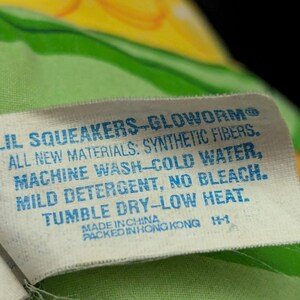 May include: A white tag with blue text reads "LIL SQUEAKERS-GLOWORM" and care instructions: "ALL NEW MATERIALS: SYNTHETIC FIBERS. MACHINE WASH-COLD WATER, MILD DETERGENT, NO BLEACH. TUMBLE DRY-LOW HEAT." The tag is on a green and yellow patterned fabric.