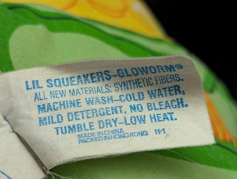 May include: A white tag with blue text reads "LIL SQUEAKERS-GLOWORM" and care instructions: "ALL NEW MATERIALS: SYNTHETIC FIBERS. MACHINE WASH-COLD WATER, MILD DETERGENT, NO BLEACH. TUMBLE DRY-LOW HEAT." The tag is on a green and yellow patterned fabric.