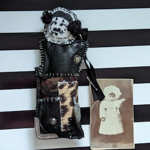 May include: A black leather doll with a white face and black hair. The doll is wearing a patchwork outfit and has a small photo of a child in a white dress attached to it.