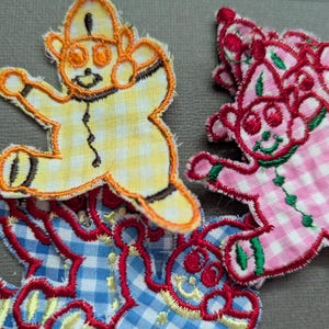 May include: Embroidered fabric patches shaped like gingerbread men. One is yellow and white gingham, another is pink and white gingham, and a third is blue and white gingham. Each patch has a smiling face and is outlined in a contrasting colour.
