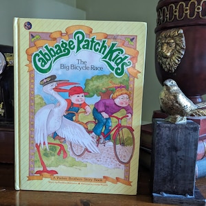 May include: A yellow book titled "Cabbage Patch Kids The Big Bicycle Race" featuring an illustration of three children riding bicycles with a white stork. The book is a Parker Brothers Story Book.