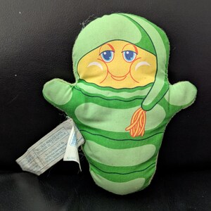 May include: A vintage plush toy in the shape of a smiling character with a yellow face, blue eyes, and a green hat and scarf. The body is green with horizontal stripes. A white tag is attached to the toy.