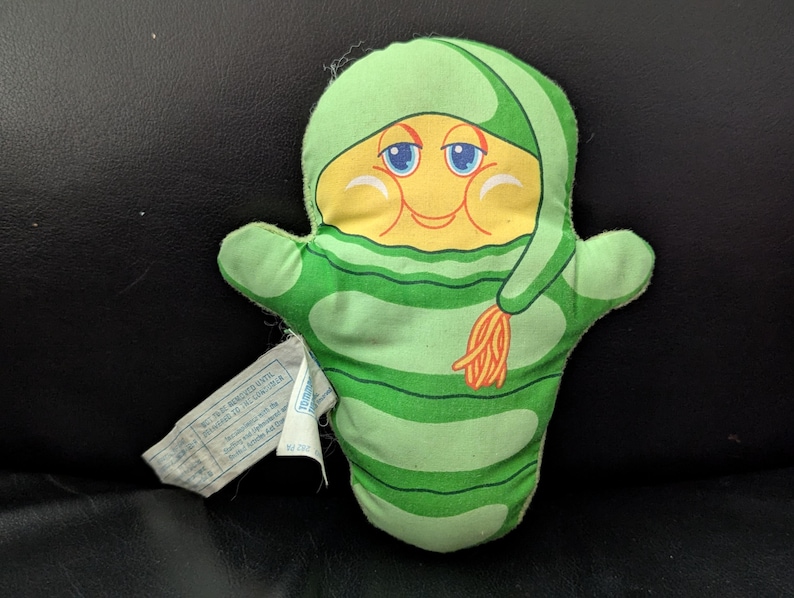 May include: A vintage plush toy in the shape of a smiling character with a yellow face, blue eyes, and a green hat and scarf. The body is green with horizontal stripes. A white tag is attached to the toy.