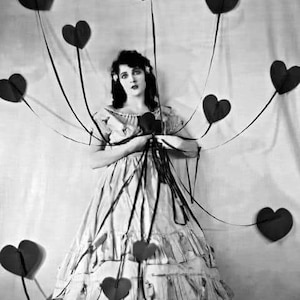 May include: A woman in a white dress stands in front of a white background. She is surrounded by black paper hearts attached to strings. The hearts are arranged in a circular pattern around her.