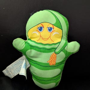 May include: A vintage plush toy in the shape of a smiling character. The toy features a yellow face with blue eyes and rosy cheeks, and is wearing a green hood and body with horizontal stripes. A tag is attached to the side.