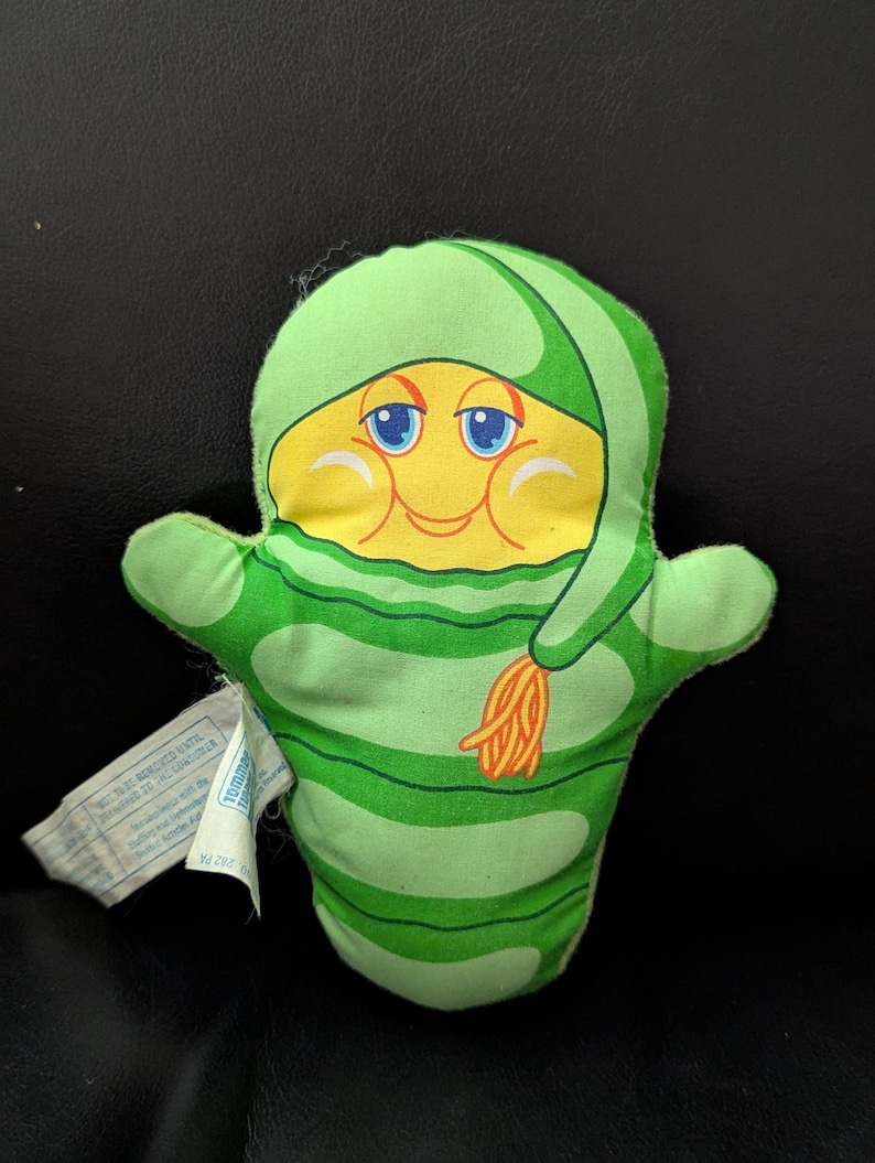May include: A vintage plush toy in the shape of a smiling character. The toy features a yellow face with blue eyes and rosy cheeks, and is wearing a green hood and body with horizontal stripes. A tag is attached to the side.