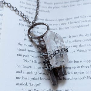 May include: A handcrafted necklace featuring a vintage key pendant adorned with a clear quartz crystal. The crystal is wrapped in silver-colored wire, suspended from a chain. The necklace rests on a book page.
