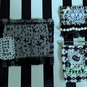 May include: Three textile art pieces featuring black, white, and gray fabrics. One rectangular patch displays the text 'AND THE SKY IS GRAY' with bird motifs. Another square patch shows a cat illustration with pearls. A third piece is a pin with a cat.
