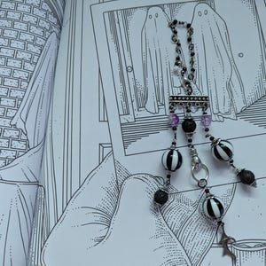 May include: A silver-toned necklace with black and white striped beads, black lava beads, and purple accents. The necklace is displayed over a black and white illustration of two ghosts.