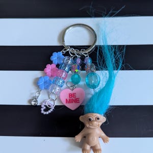May include: A blue troll keychain with a pink heart charm that says "Be Mine". The keychain also has blue and pink flower charms, clear and iridescent beads, and a silver key ring.