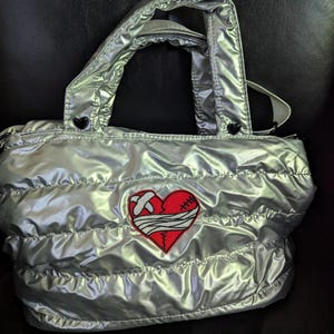 Puede incluir: Bolso tote plateado acolchado con un parche de corazón rojo. El corazón está cosido con hilo blanco y tiene un diseño de vendaje. El bolso tiene una asa superior y un cierre de cremallera. El bolso mide aproximadamente 30 cm de ancho y 25 cm de alto.