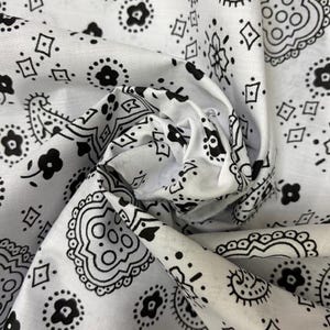 May include: A white fabric featuring a repeating black paisley and floral pattern. The material is gathered in the center, creating folds and shadows that highlight the intricate, high-contrast design of the printed shapes across the surface.
