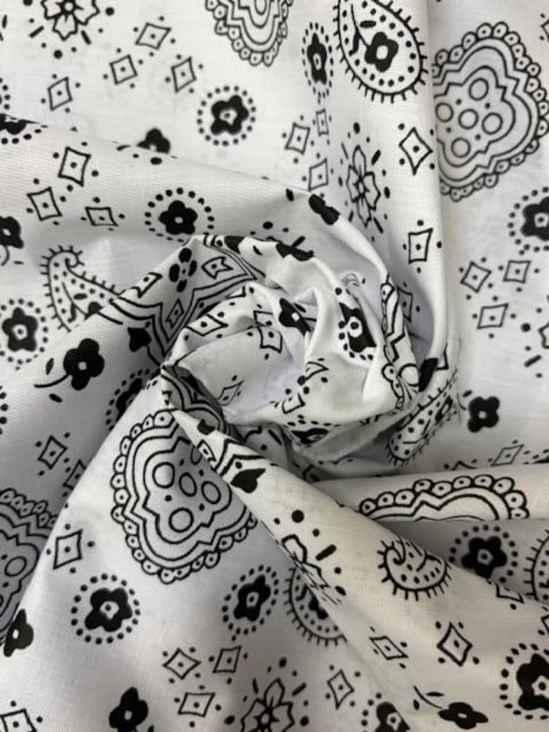 May include: A white fabric featuring a repeating black paisley and floral pattern. The material is gathered in the center, creating folds and shadows that highlight the intricate, high-contrast design of the printed shapes across the surface.