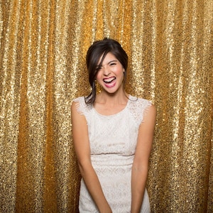 May include: A woman wearing a white lace dress smiles and laughs in front of a gold sequin backdrop.