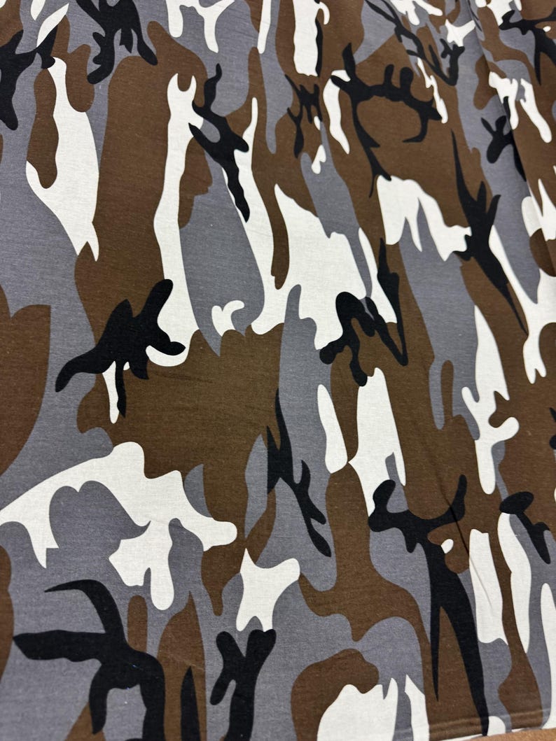 May include: Camouflage fabric with a brown, gray, and white pattern. The pattern is abstract and resembles a woodland scene.