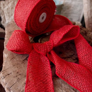May include: A roll of bright red burlap ribbon with a bow tied in the foreground. The ribbon has a coarse, woven texture and is displayed on a rustic wooden surface. The ribbon is suitable for crafts and decorations.