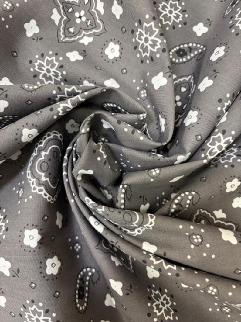 May include: A grey fabric featuring a repeating white bandana print with paisley motifs, small floral shapes, and geometric accents. The material is gathered in soft folds, highlighting the intricate pattern and the matte texture of the textile.