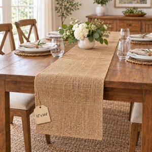 May include: A rectangular tan burlap table runner with a coarse woven texture. A beige paper tag is attached to the corner, clearly printed with the dimensions 12" x 144". The runner is laid flat across a wooden dining table with place settings.