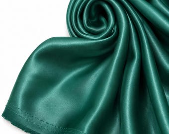 May include: A length of deep emerald green satin fabric with a smooth, lustrous finish. The material is draped in a swirling, circular pattern, highlighting the reflective quality of the textile and the visible stitched hem along the edge.