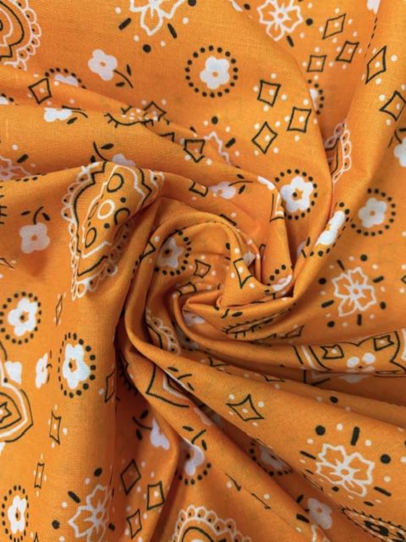 May include: A vibrant orange fabric featuring a repeating bandana pattern in white and black. The design consists of small white flowers, black diamond shapes, and intricate paisley-inspired outlines printed across the textured cotton material.