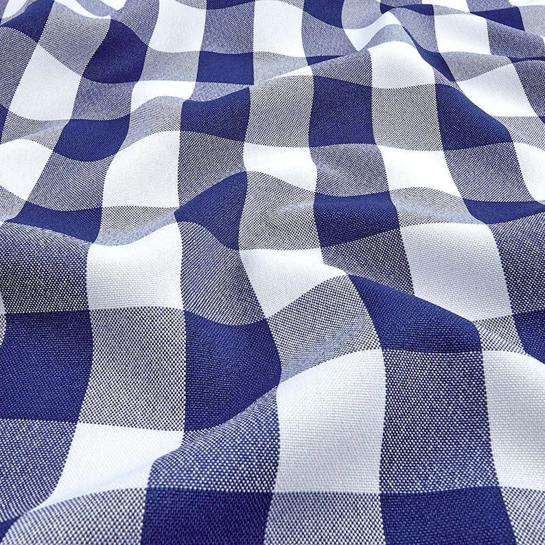 May include: Close-up of a blue and white checkered fabric. The fabric is folded and draped, creating shadows and depth. The pattern is a classic gingham design, with large, evenly sized squares. The fabric appears to be cotton or a similar material.