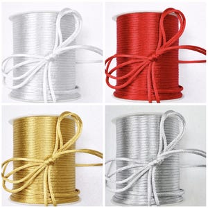 May include: Four spools of satin cord in white, red, gold, and silver. Each spool is tied with a matching satin ribbon bow. The cords are tightly wound on white plastic spools, creating a neat and organized presentation.