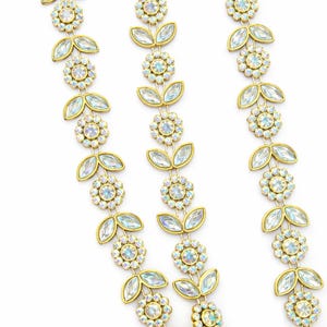 May include: Gold-toned jewelry featuring a floral and leaf design. The pieces are adorned with iridescent, clear crystals. The floral elements are centered with a cluster of crystals, and the leaf elements are elongated and pointed.