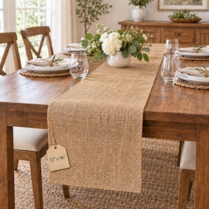May include: A rectangular tan burlap table runner with a woven texture, draped across a wooden dining table. A beige paper tag attached to the runner displays the text 12" x 96". The runner features a coarse, natural fiber weave pattern.