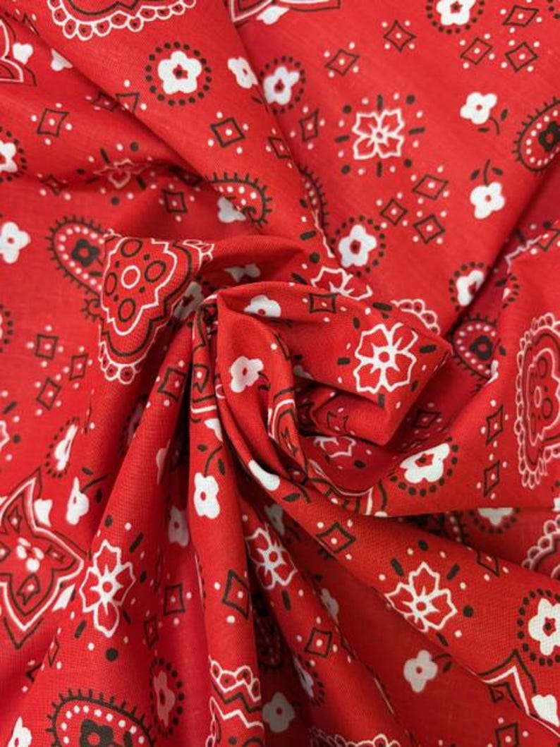 May include: A vibrant red fabric featuring a classic bandana print with repeating white floral motifs, black paisley shapes, and small diamond outlines. The material is gathered in the center, creating soft folds that highlight the intricate pattern.