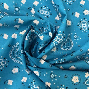 May include: A bright turquoise fabric featuring a repeating white and black bandana print. The pattern consists of stylized floral motifs, small white blossoms, and intricate paisley shapes arranged across the surface of the folded textile.