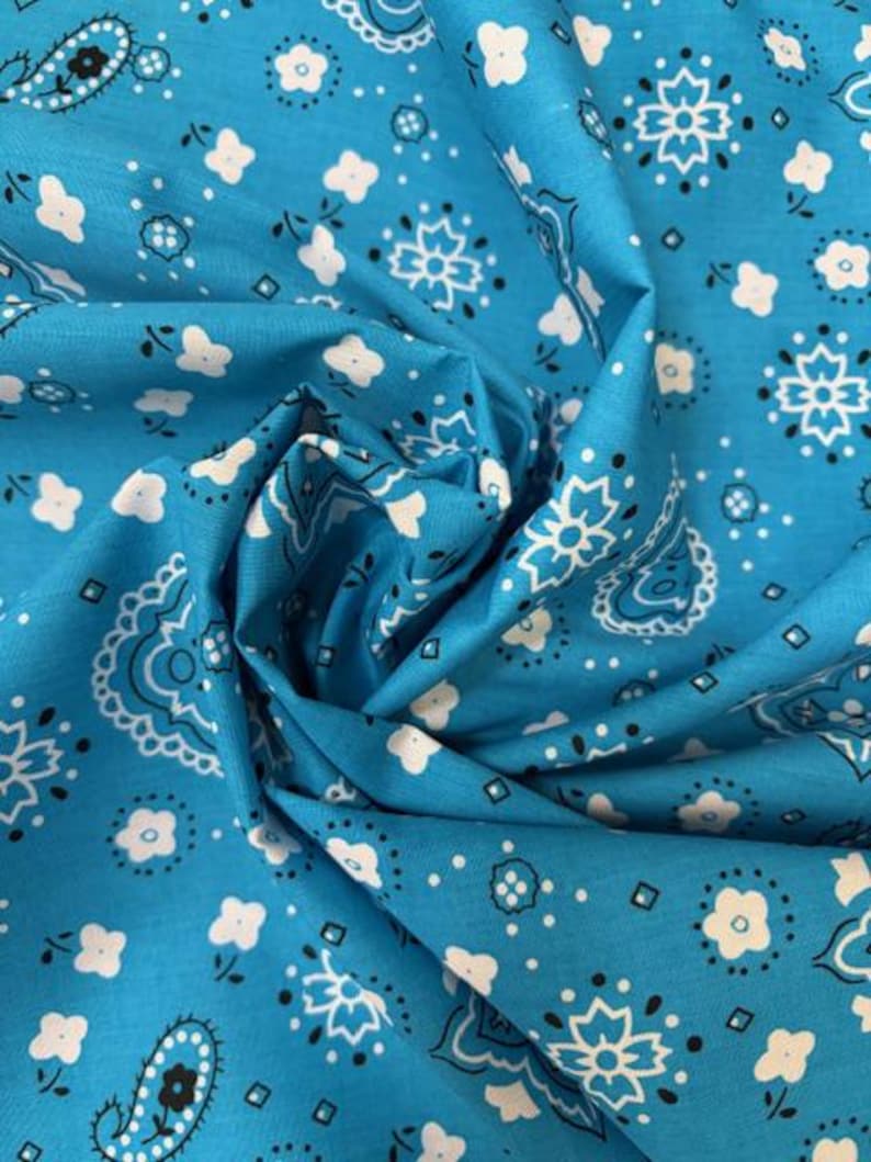 May include: A bright turquoise fabric featuring a repeating white and black bandana print. The pattern consists of stylized floral motifs, small white blossoms, and intricate paisley shapes arranged across the surface of the folded textile.