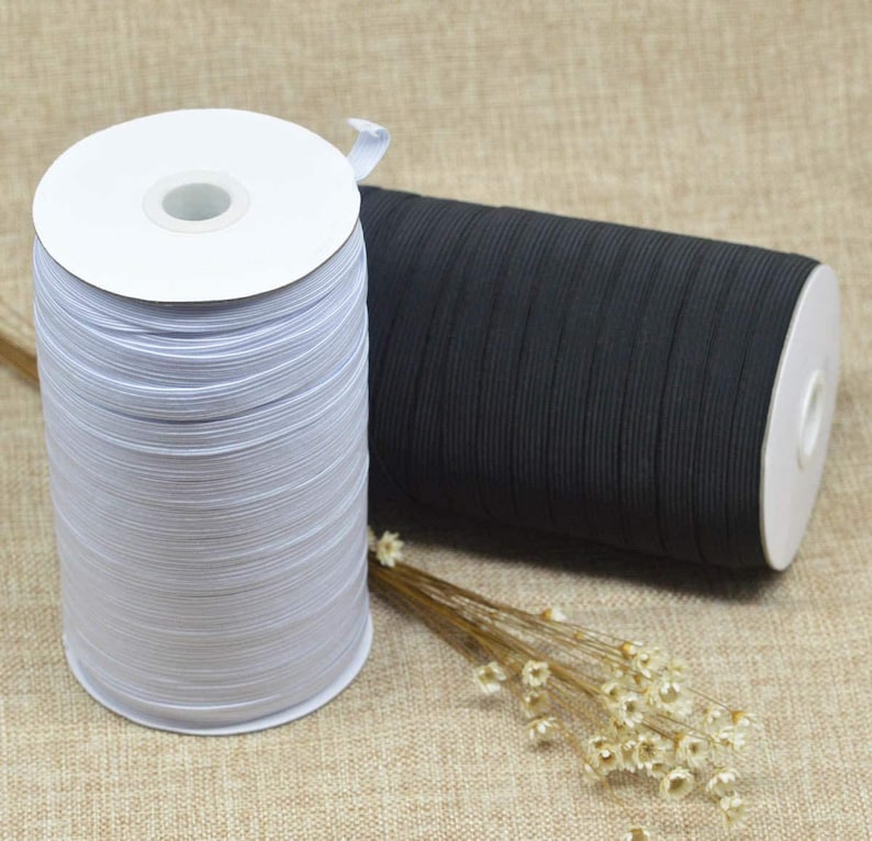 May include: Two spools of elastic cord, one white and one black, are wound around cardboard spools. The spools are sitting on a brown fabric surface.