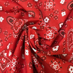 May include: A vibrant red fabric featuring a classic bandana print with repeating white floral motifs, black paisley shapes, and small diamond outlines. The material is gathered in the center, creating soft folds that highlight the intricate pattern.
