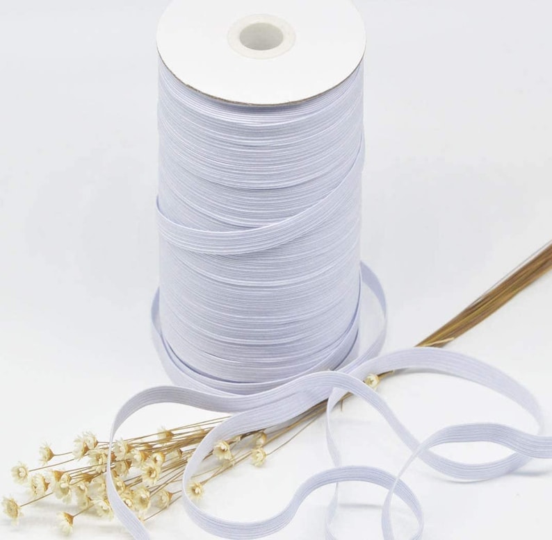May include: A spool of white elastic cord, wound around a cardboard tube. The elastic cord is lying on a table with a sprig of dried flowers.