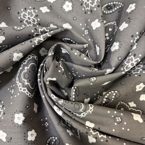 May include: A grey fabric featuring a repeating white bandana print with paisley motifs, small floral shapes, and geometric accents. The material is gathered in soft folds, highlighting the intricate pattern and the matte texture of the textile.
