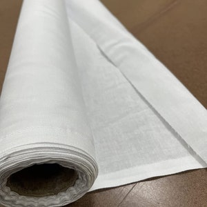 May include: A white fabric roll, unfurled on a brown surface. The fabric is smooth and appears to be a lightweight cotton.
