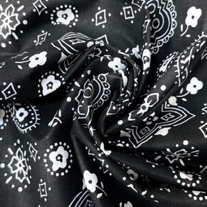 May include: A black fabric featuring a repeating white pattern of small flowers, geometric diamond shapes, and intricate paisley-inspired swirls. The material is gathered in the center, creating soft folds and shadows across the surface.