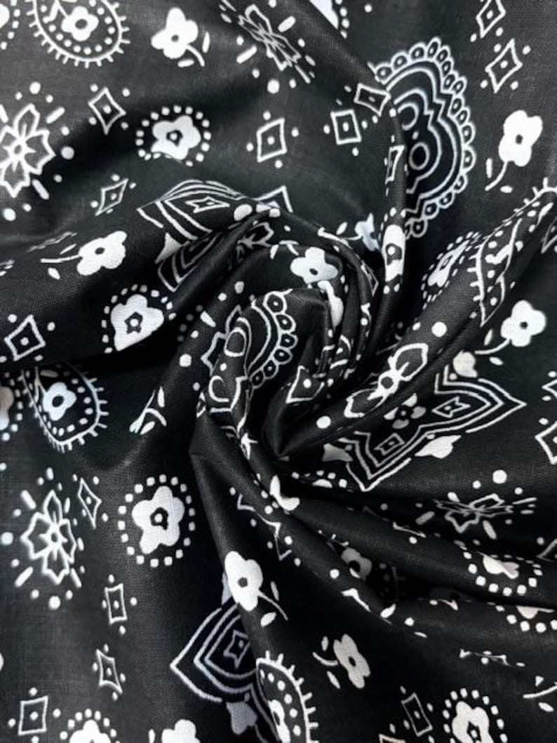 May include: A black fabric featuring a repeating white pattern of small flowers, geometric diamond shapes, and intricate paisley-inspired swirls. The material is gathered in the center, creating soft folds and shadows across the surface.