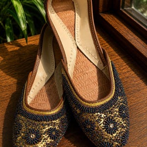 May include: A pair of slip-on flats featuring a brown leather trim and a gold-toned base. The exterior is heavily embellished with dark blue beads and gold sequins arranged in a floral pattern. The interior sole has a textured, wave-like pattern.
