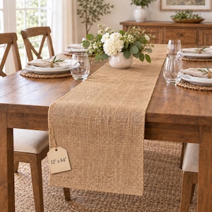 May include: A rectangular tan burlap table runner with a loose-weave texture, featuring a beige paper tag printed with the dimensions 12" x 84". The runner is laid flat across a wooden dining table, centered beneath a white floral centerpiece.