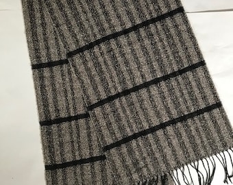 May include: A rectangular scarf with vertical stripes in shades of gray and brown, accented by three horizontal black stripes. The scarf has a fringed edge. This accessory is suitable for cold weather.