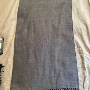 May include: A rectangular, gray scarf with a fringed edge. The scarf is made of a soft, woven material and is displayed on a neutral-colored surface. The fringe is a darker shade of gray.