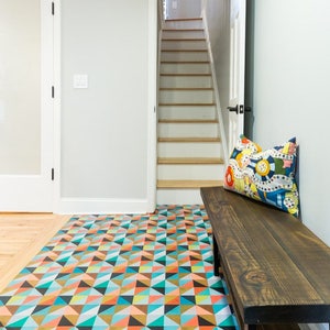 May include: A wooden bench with a colorful geometric patterned rug underneath. The bench has a black metal frame and a dark brown wooden seat. A colorful pillow with a cartoon design is on the bench. The bench is in front of a doorway leading to a staircase.