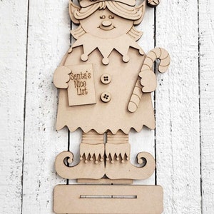 May include: Wooden elf cutout with a candy cane, a Santa's Nice List sign, and buttons. The elf is standing on a wooden platform.