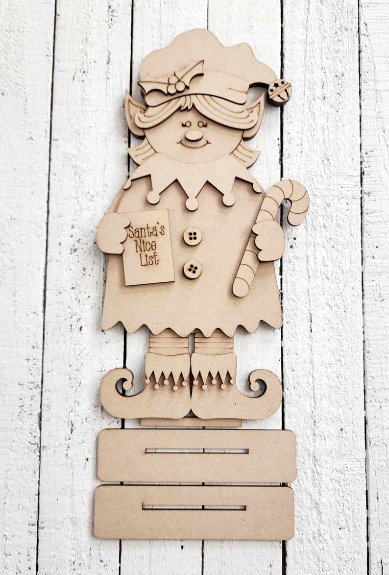 May include: Wooden elf cutout with a candy cane, a Santa's Nice List sign, and buttons. The elf is standing on a wooden platform.