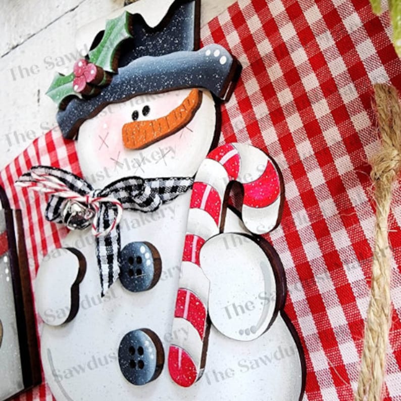 May include: A wooden snowman ornament with a black top hat, red and white candy cane, and a black and white plaid scarf. The snowman is painted white with black buttons and a red nose. The ornament is on a red and white checkered background.