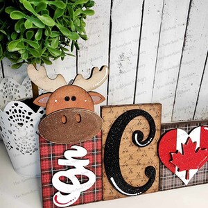 May include: Wooden sign with the word "Camp" spelled out in black letters with glitter. The "C" is a large block letter and the "amp" is in smaller letters. The "C" is decorated with a pattern of camping axes and tents. The sign is decorated with a red and black plaid pattern, a moose head, and a red heart with a Canadian flag.