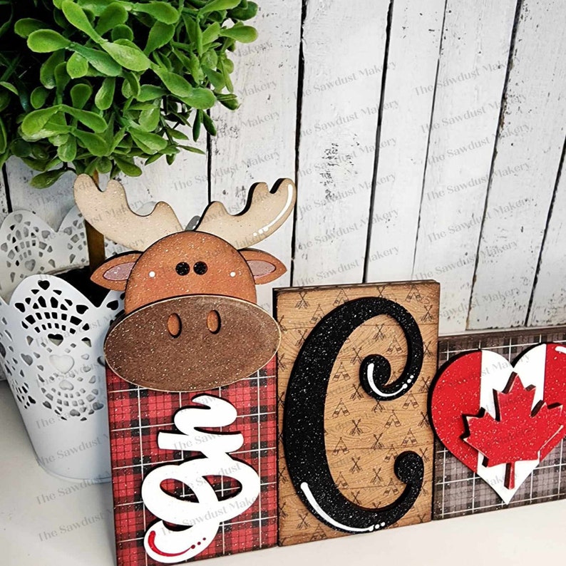May include: Wooden sign with the word "Camp" spelled out in black letters with glitter. The "C" is a large block letter and the "amp" is in smaller letters. The "C" is decorated with a pattern of camping axes and tents. The sign is decorated with a red and black plaid pattern, a moose head, and a red heart with a Canadian flag.