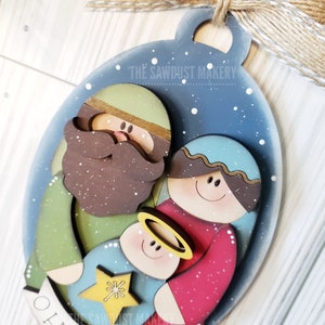 May include: A wooden Christmas ornament depicting the nativity scene with Joseph, Mary, and baby Jesus. The ornament is painted in a blue, green, red, and brown color scheme and features a white star and the text "O Holy Night".